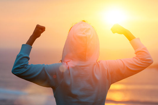 A person making a 'strength' pose wearing a hoodie next to the ocean at sunrise